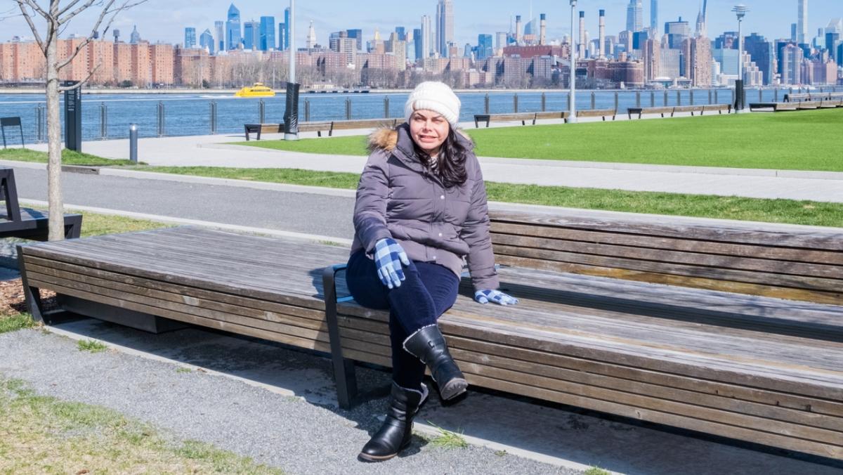 Cristina Herrera in a hat, coat, and gloves, sitting on a park bench