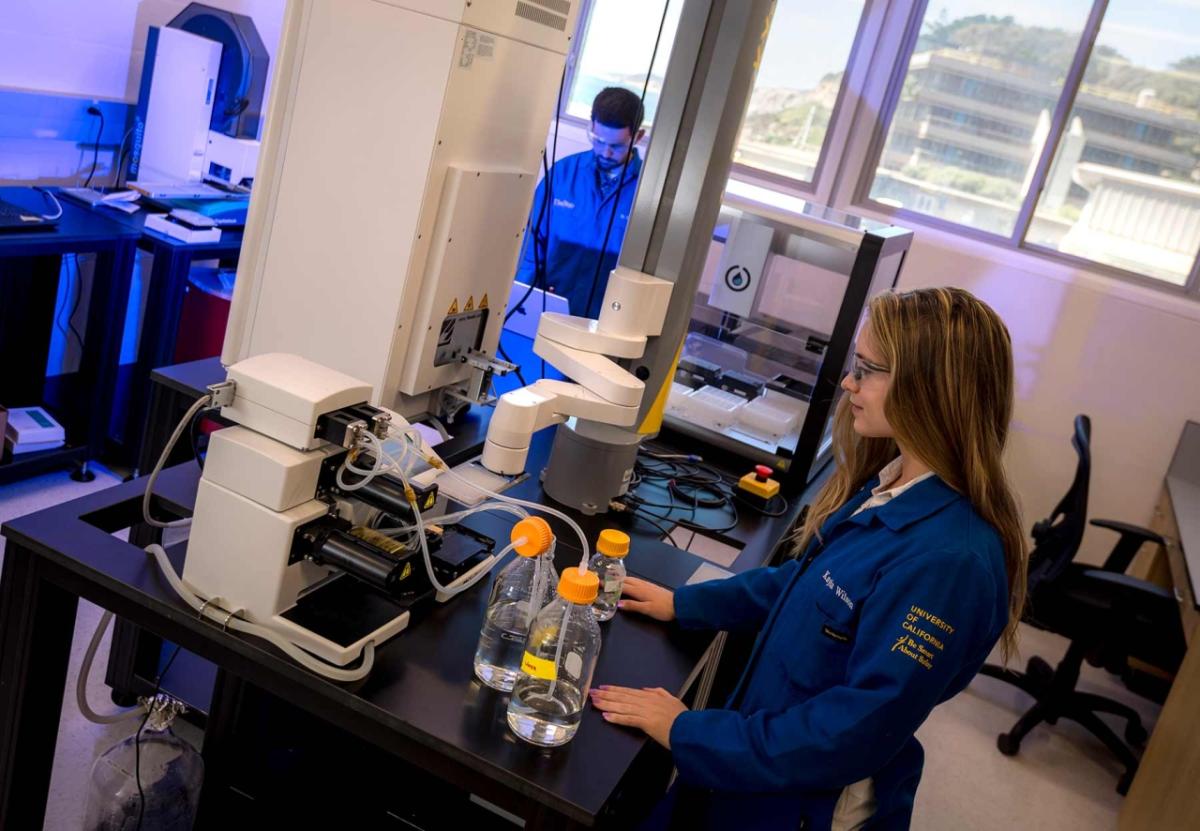 In the Illumina Lab in Scholander Hall, PhD student Kayla Wilson operates a new bulk reagent dispenser, which can rapidly fill well plates with two different liquids. Postdoctoral scholar Timothy Fallon programs a liquid handling robot.