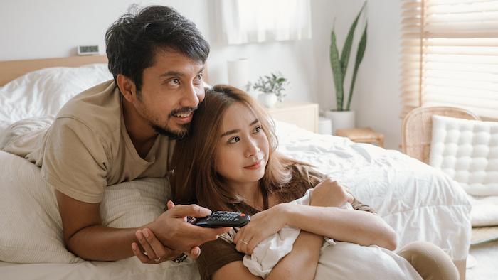 Asian couple watching television in a bedroom.