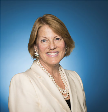 Profile of Sue Kronick. Blue background. Wearing a beige suit and pearl necklace