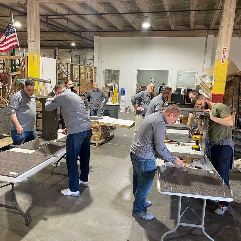 A group of volunteers assembling cabinets on long tables in a warehouse.