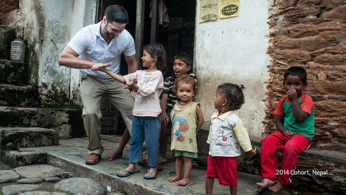 an adult in khakis, a polo shirt and flip flops shakes the hand of a child while four others look on "2014 cohort Nepal" in the corner