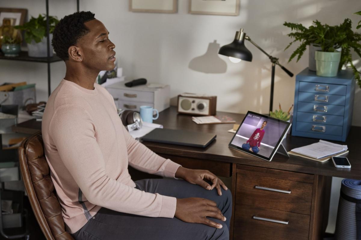 a person sitting at a home office desk, eyes closed, a meditation program playing on the device on their desk