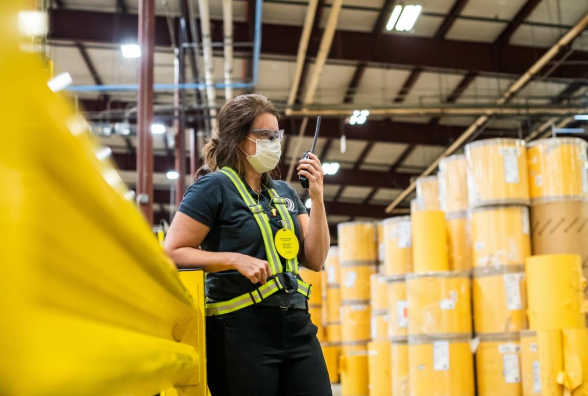 Karen Crisp in safety gear standing among yellow barrels in a factory