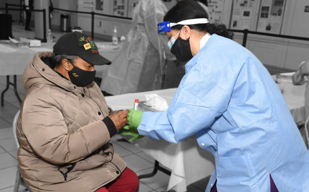 a person in protective medical apparel collects a sample in a test tube from another person seated in front of them
