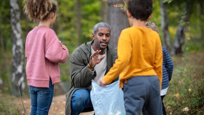 Man speaking and teaching  three children in a wooded area. 