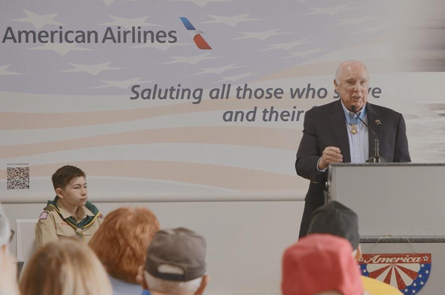 person stands at a podium in front of a crowd. American Airlines logo behind them