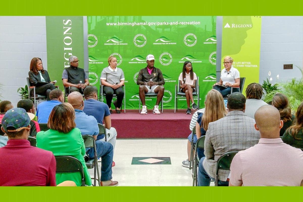 A panel of Black leaders, athletes, and students on a raised platform in front of a green backdrop