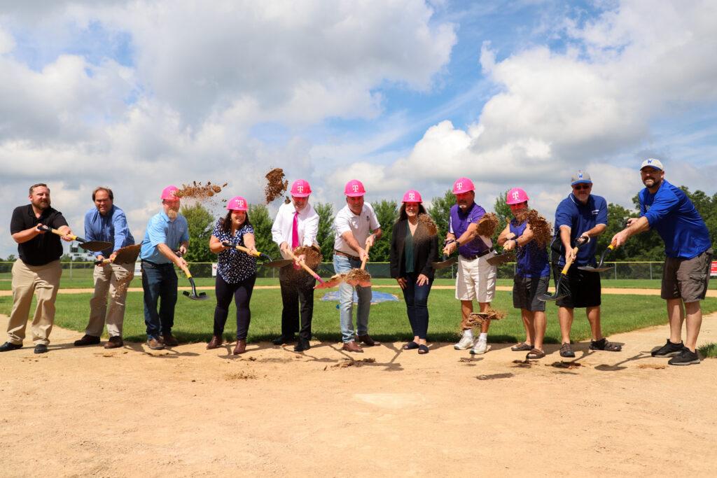 a group of people with shovels digging sand in a baseball field, all wearing T-Mobile, pink hardhats