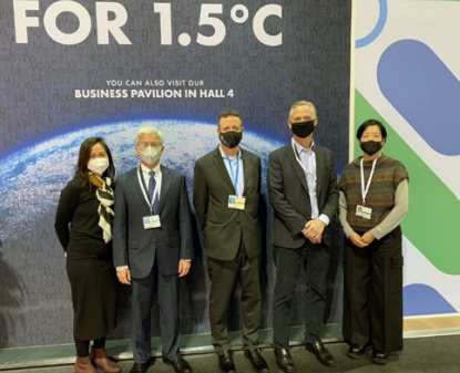 a group of five people in business attire, in front of a "For 1.5 degree C" banner. All are wearing medical masks.